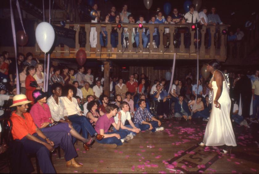 Photograph of a drag queen performing in a coral dress and white feather boa as part of the Valentine's Day drag show at theBar. The audience on the first and second floors are visible, watching the performance.