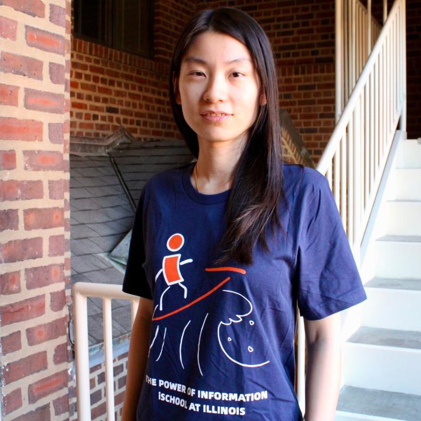 Meng Li wears the T-shirt with her winning design. The shirt is dark blue, with a hand-sketched wave in white, while the figure and surf board are in Illini Orange.