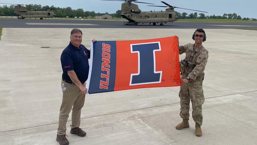 Michael Wonderlich and Michael Ferrer hold a U of I flag in front of a military helicopter