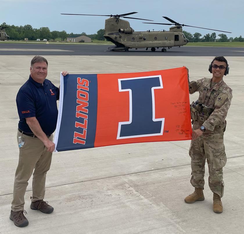 Michael Wonderlich and Michael Ferrer hold a U of I flag in front of a military helicopter